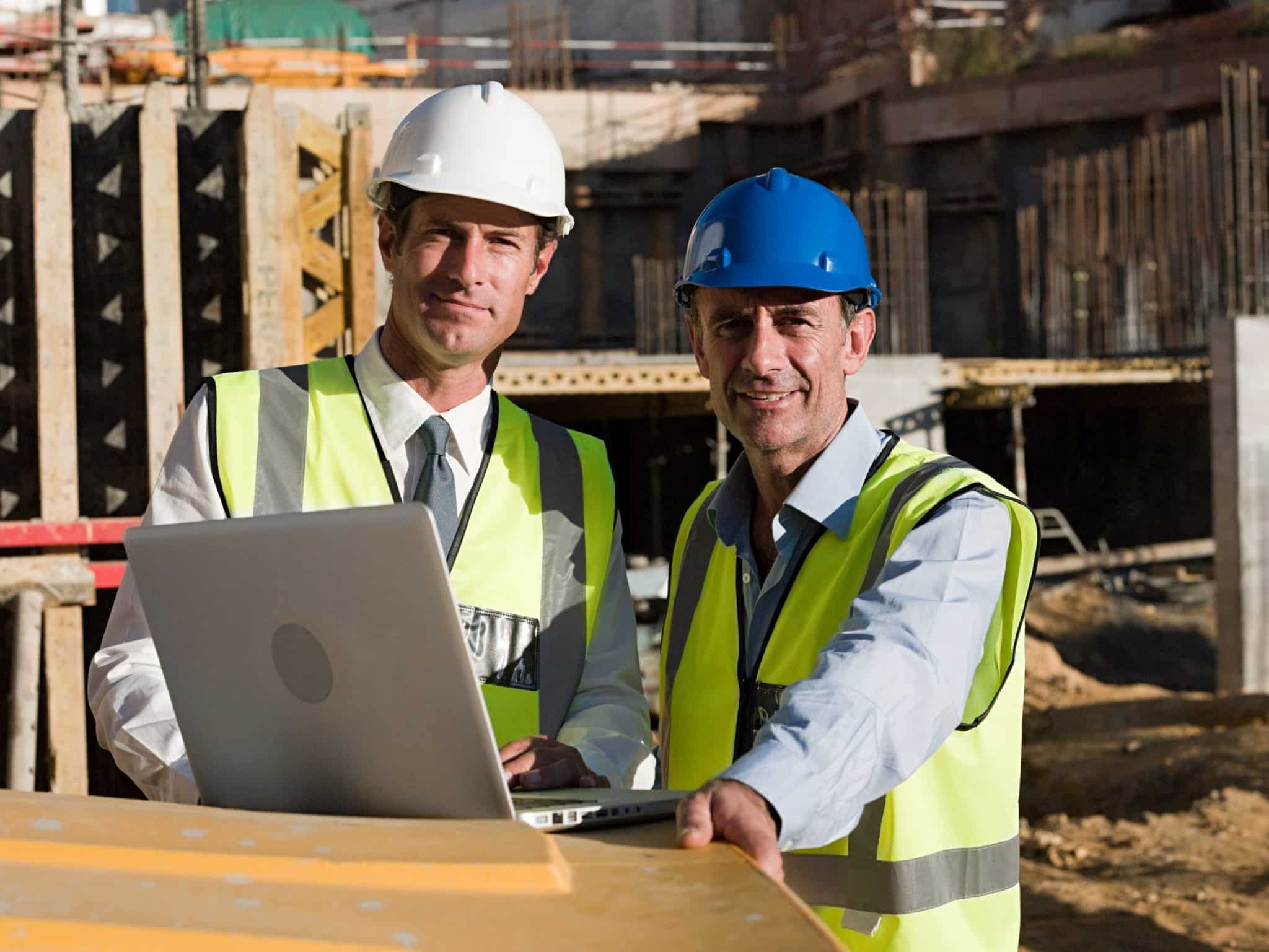 Mature men using laptop on construction site while wearing safety vests and hard hats.